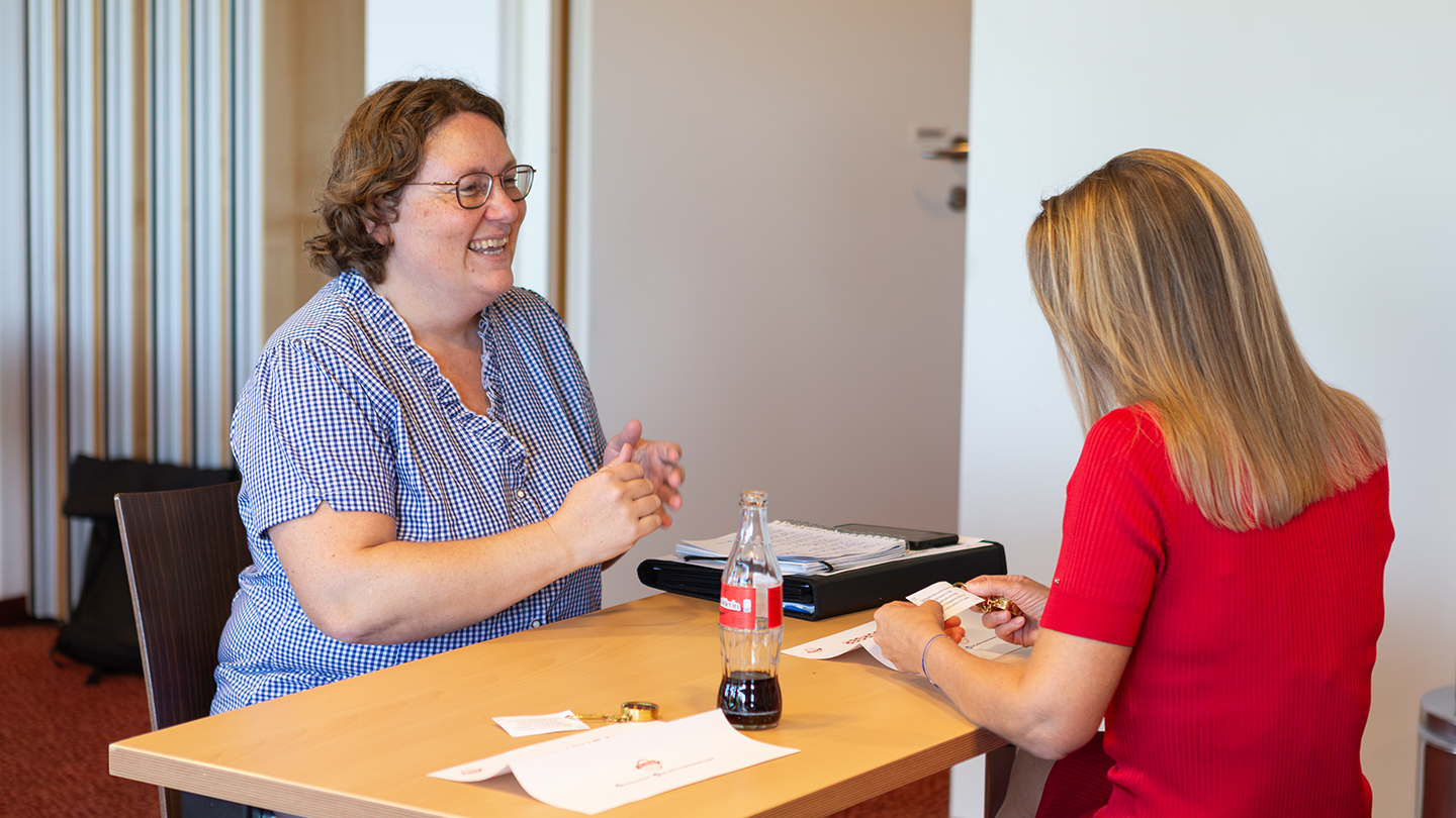 Zwei Frauen sitzen an einem Tisch und sprechen miteinander. Eine der beiden lacht herzlich, auf dem Tisch liegen Unterlagen und eine Cola-Flasche – eine Szene aus den persönlichen Coaching-Sessions des Wochenendes.