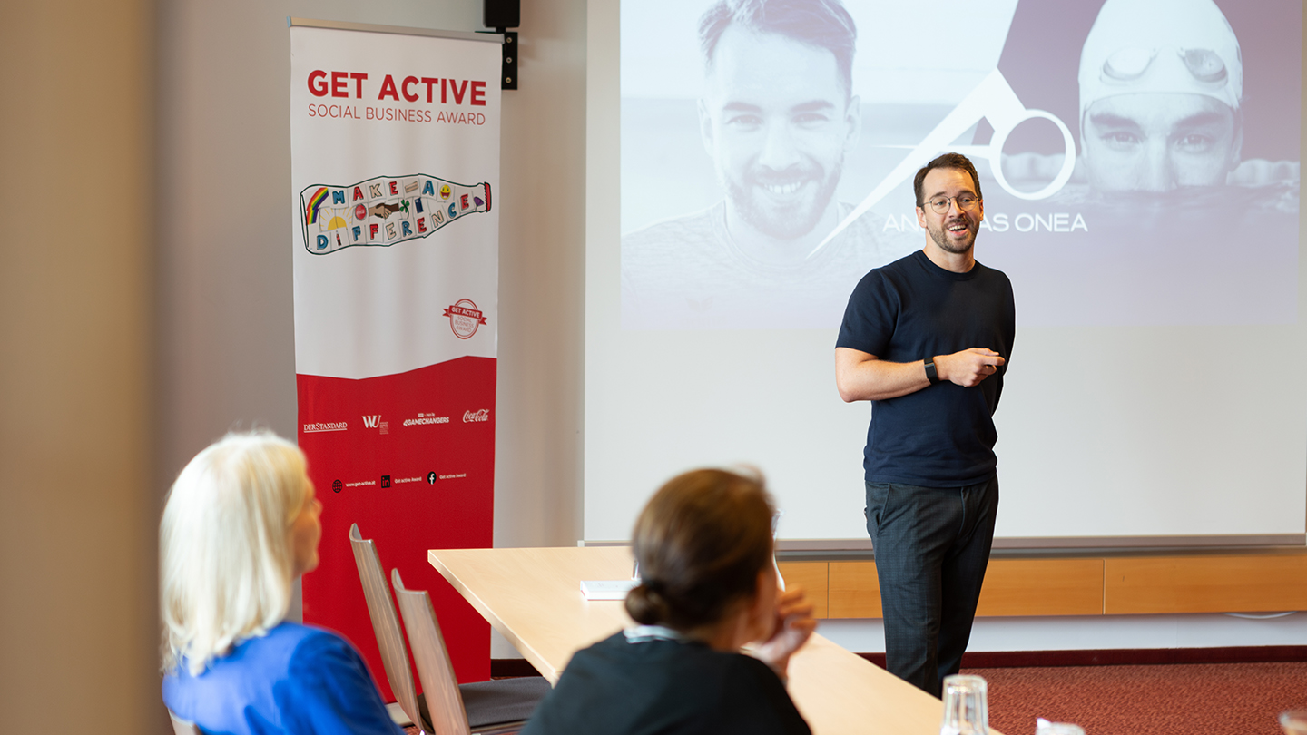 Ein Mann im dunklen T-Shirt spricht vor den Teilnehmer:innen. Auf der Leinwand hinter ihm sind Fotos von ihm als Schwimmer und Speaker zu sehen, daneben das Logo mit seinem Namen „Andreas Onea“.