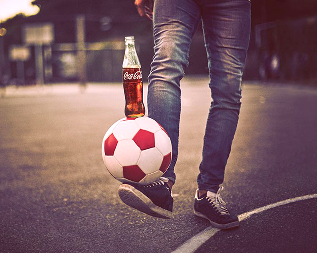 La photo représente une personne dont on ne voit que les jambes, debout sur un terrain de sport, tenant un ballon de football en équilibre sur son pied. Une bouteille en verre de Coca-Cola est posée sur le ballon.