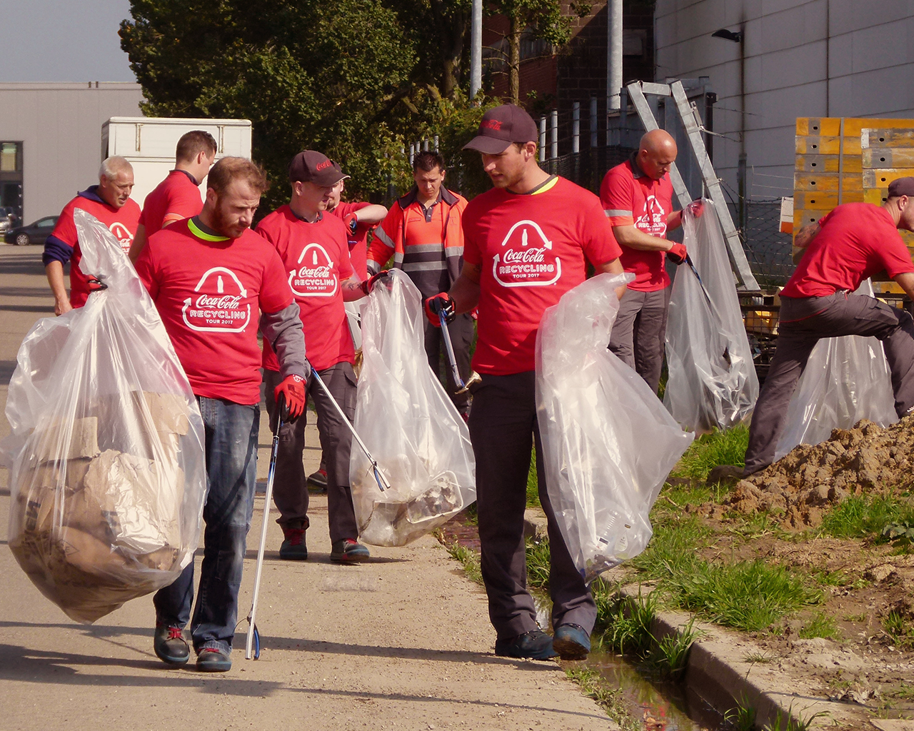 La photo représente un groupe de personnes portant des t-shirts rouges avec le logo Coca-Cola et la mention "I love Recycling". Elles participent à une collecte de déchets en extérieur, tenant de grands sacs en plastique transparents et utilisant des pinces pour ramasser les détritus le long d’un chemin et d’un terrain herbeux.