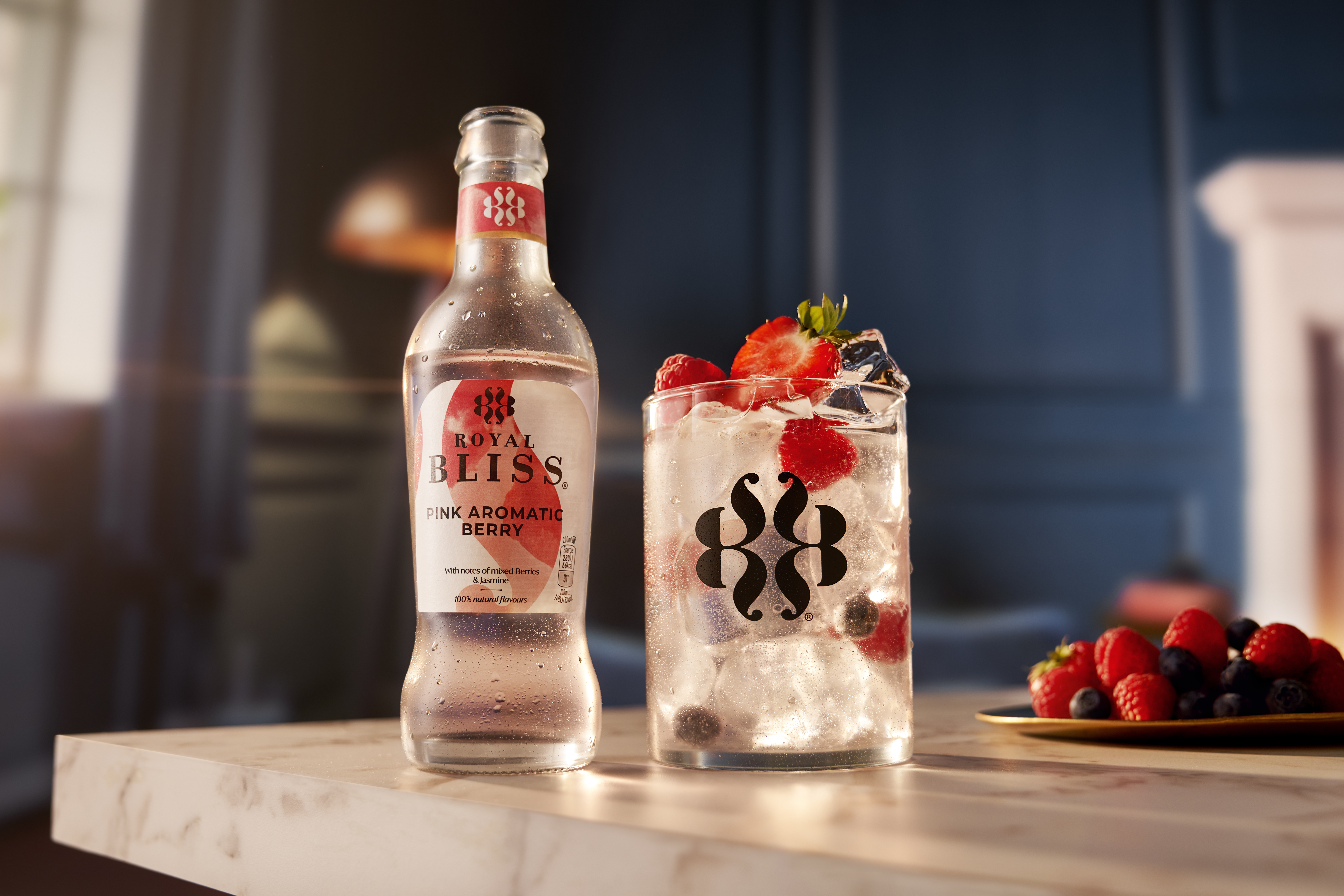 Bouteille « Royal Bliss – Pink Aromatic Berry » remplie de boisson rose clair, posée sur un plan de travail à côté d’un verre rempli de glaçons et de baies rouges et bleues entières flottant dans la boisson pétillante. En arrière-plan, ambiance chaleureuse avec assiette de fruits rouges.