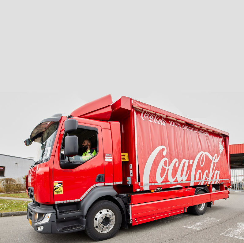 Photo avec un camion rouge Coca‑Cola 100 % électrique, garé près d’un bâtiment industriel.