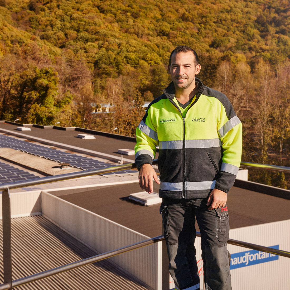 Photo avec un employé en tenue de sécurité haute visibilité sur le toit de l’usine Chaudfontaine en Belgique, avec panneaux solaires et colline boisée en arrière-plan.