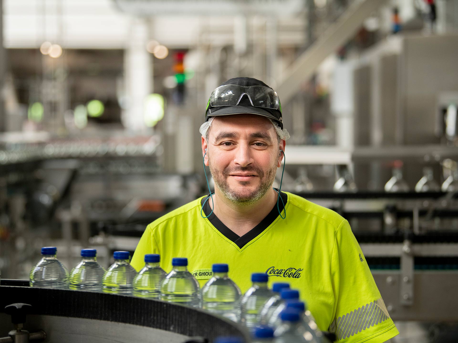 Foto met een werknemer in hoogzichtbare veiligheidskleding bij een productielijn van waterflessen.