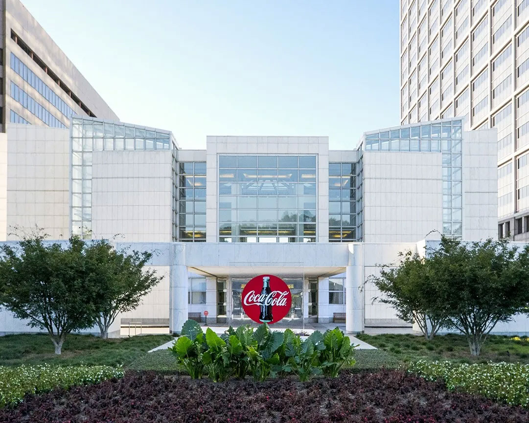 Photo avec le bâtiment moderne du siège de Coca‑Cola à Atlanta, façade blanche vitrée, logo rouge de la marque à l’entrée, entouré de végétation et d’arbres.