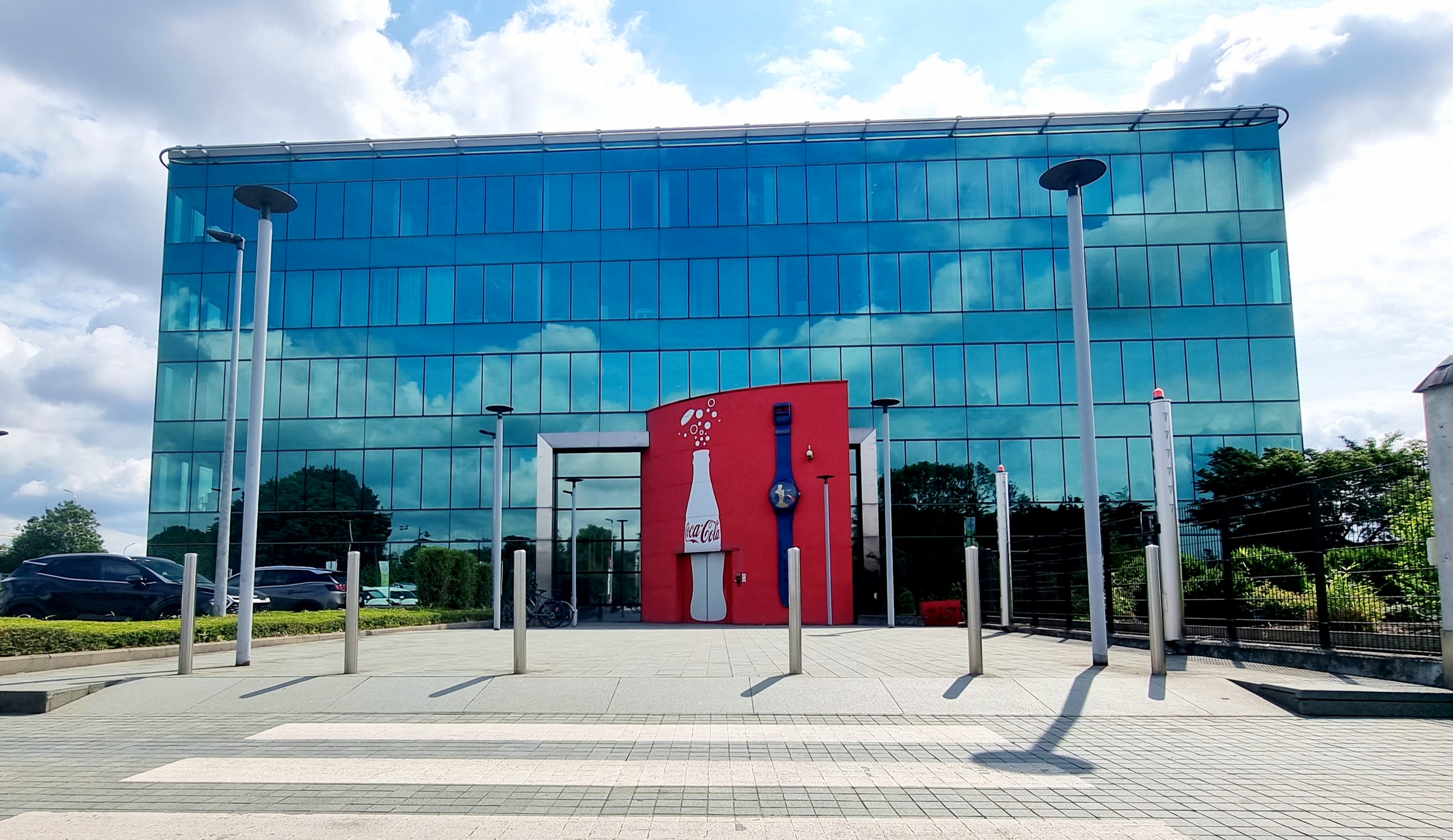 Photo avec le siège de Coca‑Cola en Belgique, bâtiment moderne à façade vitrée bleue, entrée ornée d’un mur rouge avec une bouteille blanche Coca‑Cola et une grande montre décorative.