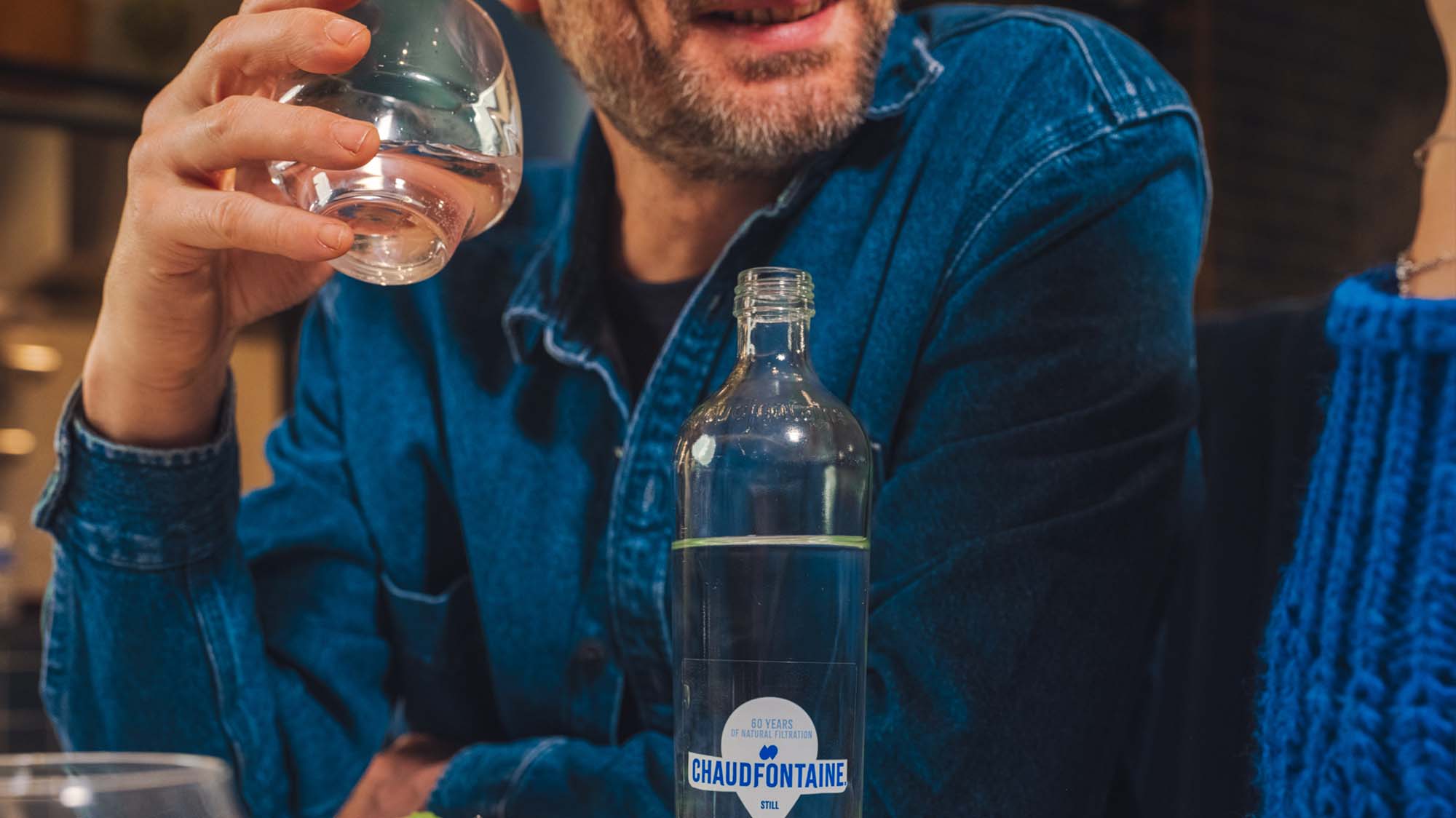 Close-up van een man in een spijkerhemd die een glas water bij zijn mond houdt, met een glazen fles Chaudfontaine op tafel. Rechts in beeld is een andere persoon te zien met een blauwe trui.