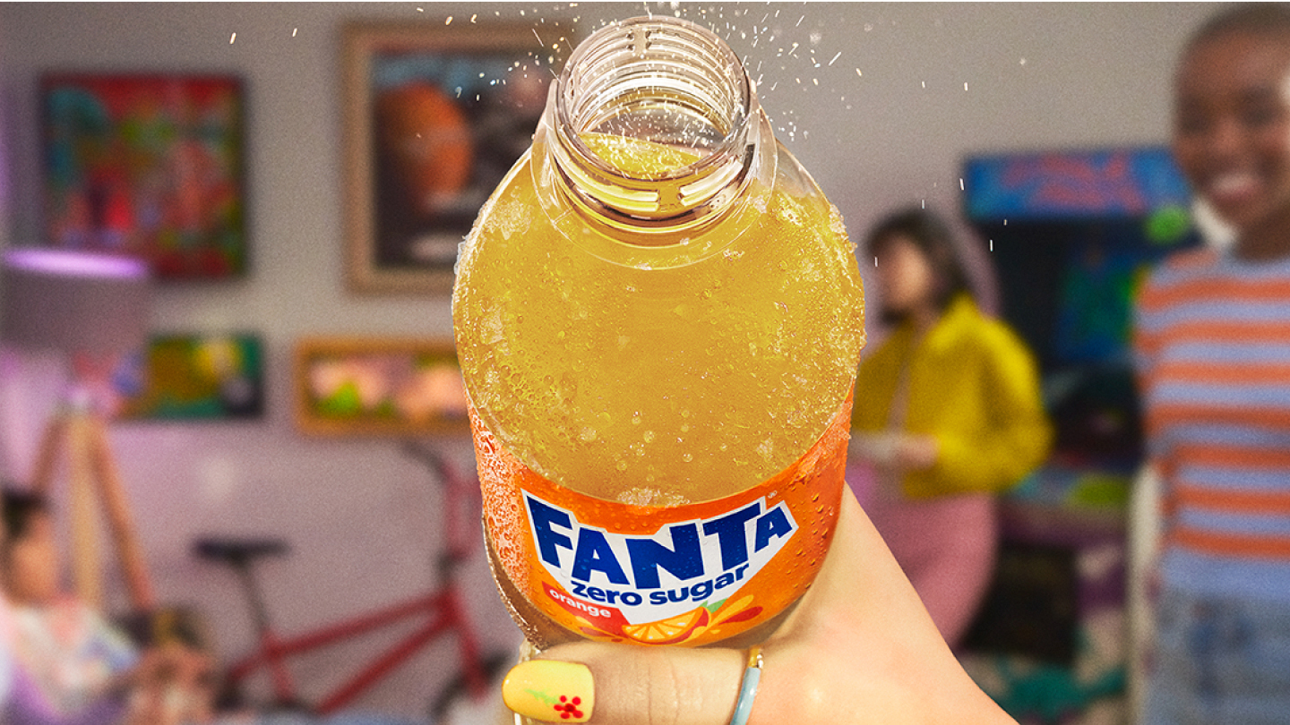 Close-up van een hand die een open fles Fanta Orange zonder suiker vasthoudt, waarbij de bruisende drank te zien is met belletjes en opspattende druppeltjes. Op de onscherpe achtergrond is een kleurrijke kamer te zien met schilderijen aan de muur, meubels en mensen die met elkaar praten.