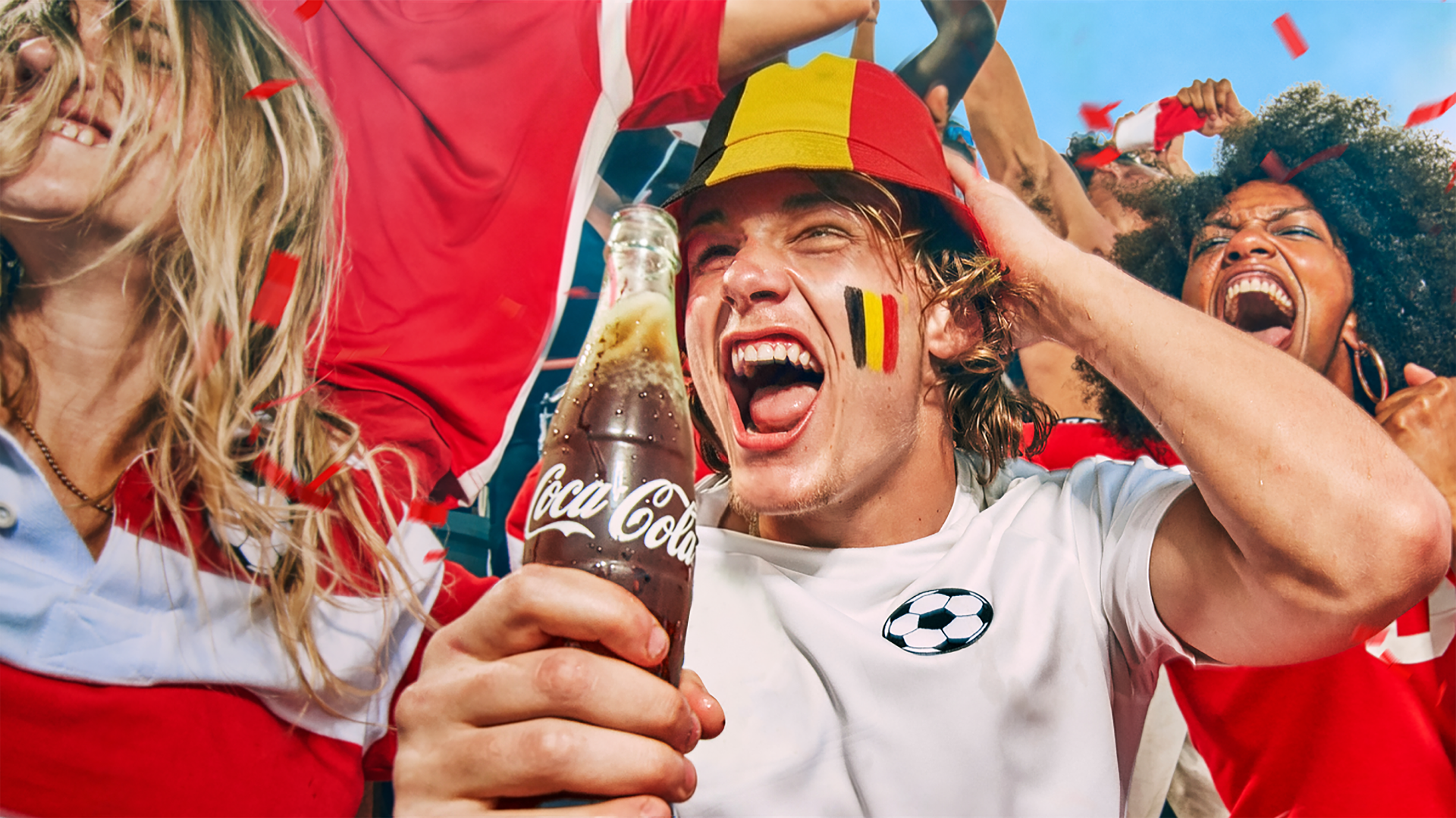 Feestelijke groep supporters in rood‑witte kleding die juichen; op de voorgrond wordt een gekoelde Coca‑Cola‑fles omhoog gehouden terwijl confetti door de lucht valt, met een zomerse sfeer en voetbalbeleving.