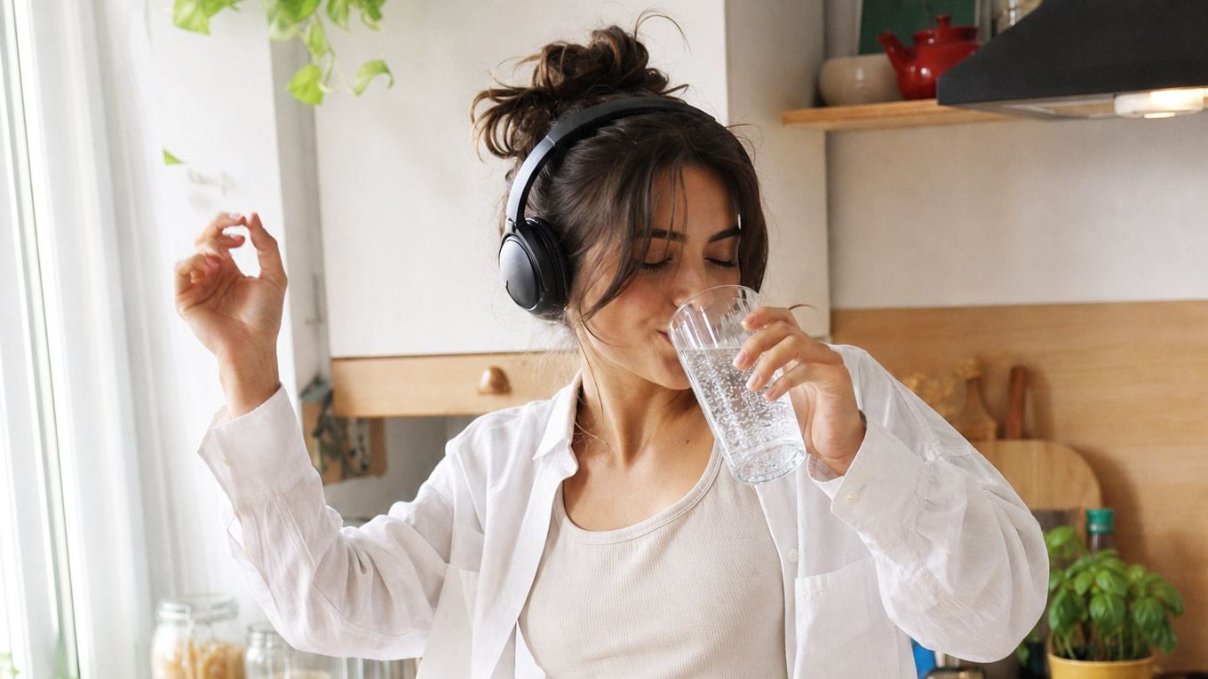 Jeune femme en tenue blanche et casque audio circum-aural boit dans un grand verre d'eau dans la cuisine