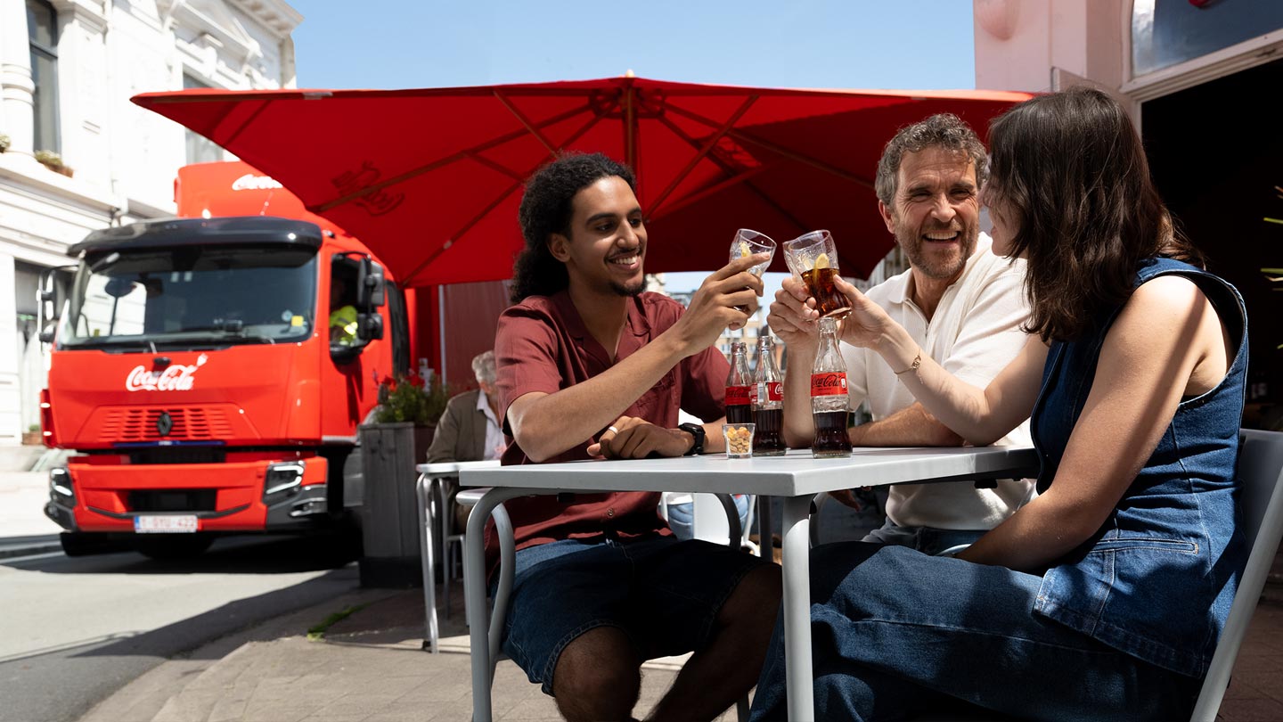 Trois personnes assises à une table en extérieur trinquent avec des verres de Coca-Cola, tandis que des bouteilles de la marque sont posées sur la table. En arrière-plan, un grand parasol rouge et un camion rouge sont visibles dans une rue ensoleillée.