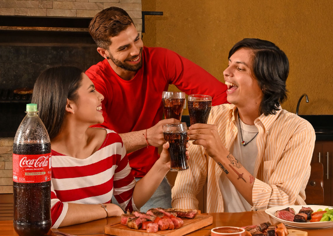 Tres jóvenes ríen y brindan con Coca-Cola Sabor Original en una parrillada.