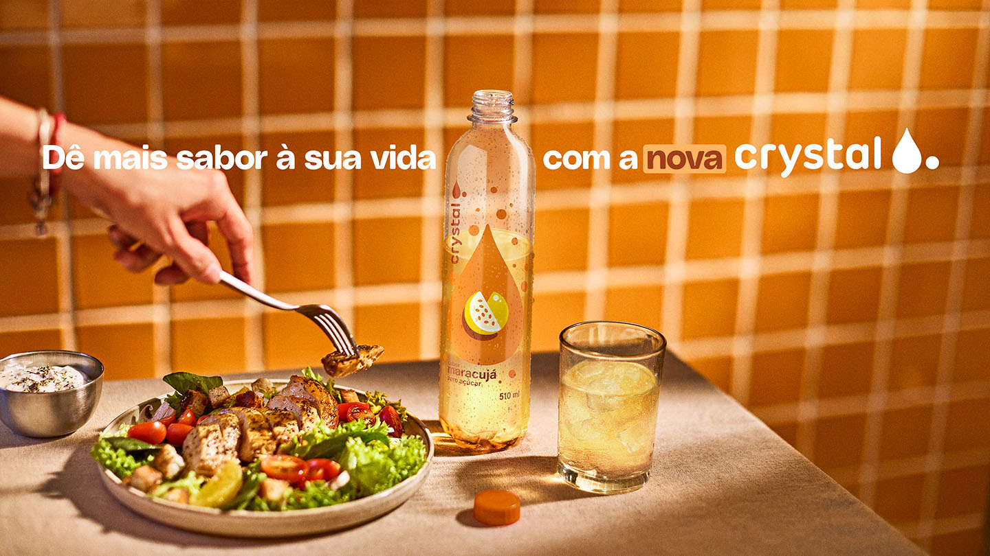 Refeição saudável com salada e frango grelhado, acompanhada pela nova água saborizada Crystal de Maracujá. A garrafa e um copo com a bebida estão sobre a mesa, destacando a mensagem 'Dê mais sabor à sua vida com a nova Crystal'.