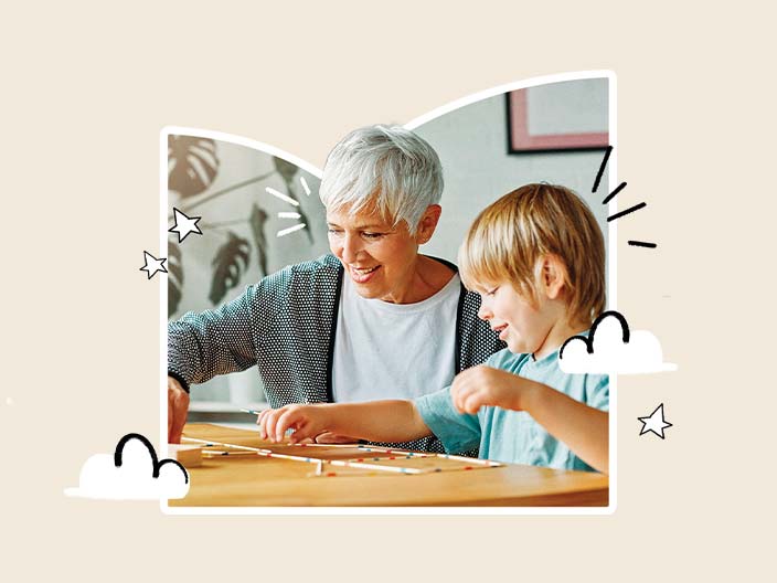 Uma avó sorridente e seu neto brincam juntos em uma mesa com palitos e blocos de madeira. A imagem transmite alegria e conexão familiar.