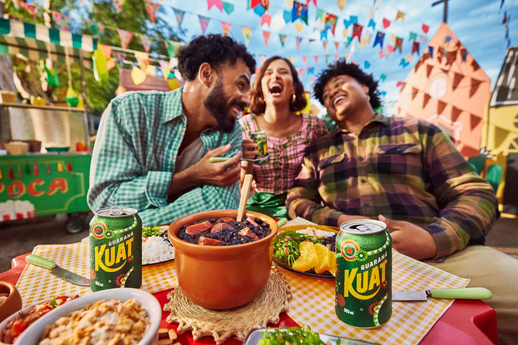 Três amigos sorrindo em uma Festa, com Guaraná Kuat, feijoada e outros quitutes em uma mesa xadrez, com bandeirinhas e decoração festiva ao fundo.