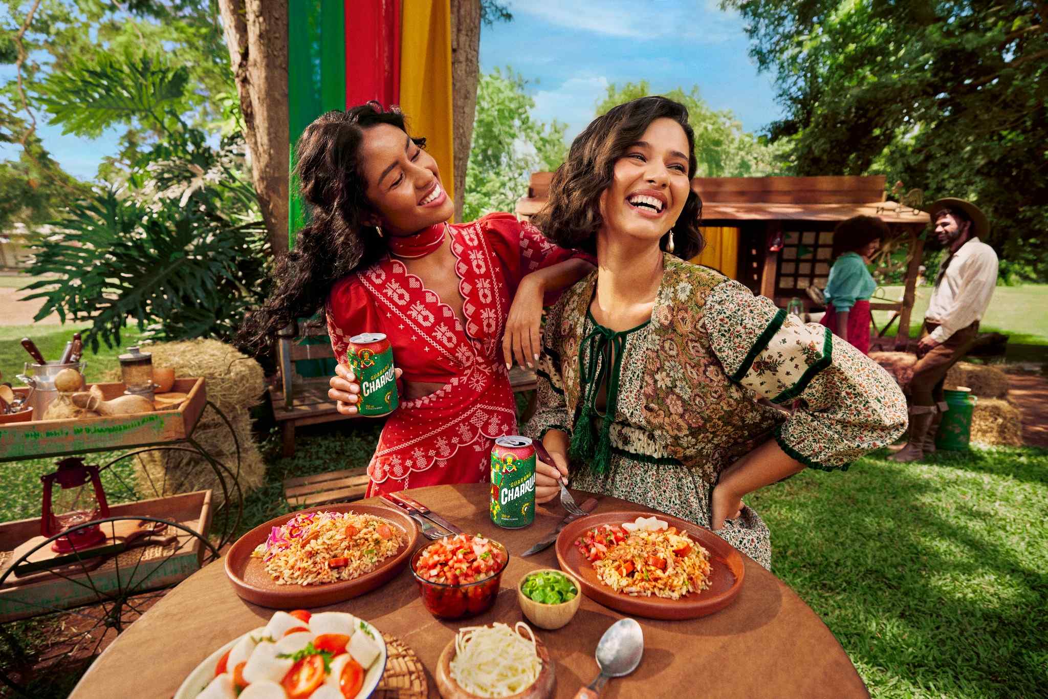 Duas amigas sorrindo e celebrando com Guaraná Charrua em um piquenique ao ar livre. Comida típica e paisagem campestre ao fundo.