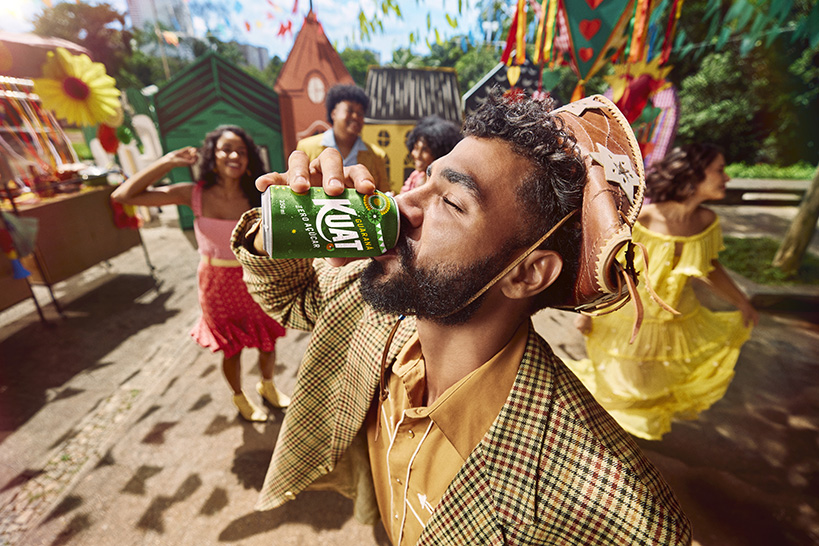 Três amigos sorrindo em uma Festa, com Guaraná Kuat, feijoada e outros quitutes em uma mesa xadrez, com bandeirinhas e decoração festiva ao fundo.