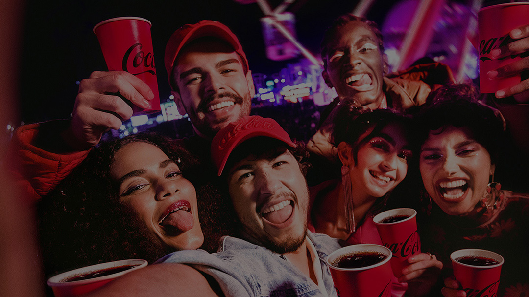 Grupo de jovens sorrindo e se divertindo em um festival à noite, segurando copos vermelhos da Coca-Cola. O ambiente está iluminado com luzes vibrantes, criando uma atmosfera festiva.