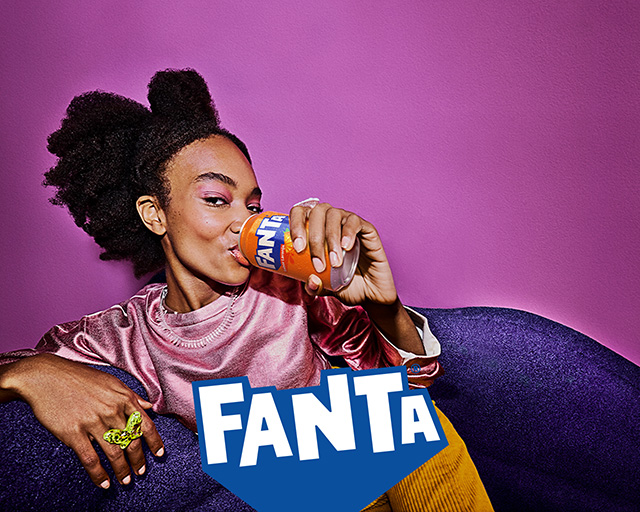 Uma mulher está sentada em um sofá roxo, bebendo uma lata de Fanta. Ela está vestindo uma blusa rosa e calças amarelas. O fundo é uma parede rosa. O logotipo da Fanta está visível na frente do sofá