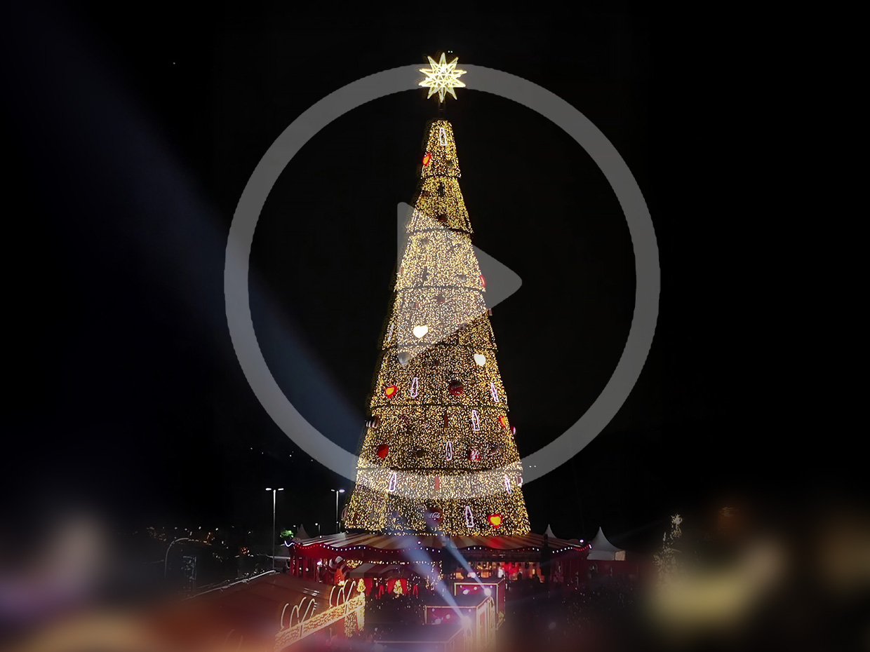 Uma grande árvore de Natal iluminada à noite, com uma estrela no topo e um ícone de "play" sobreposto.