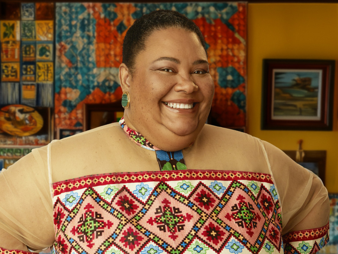 Uma mulher sorrindo, usando um vestido com estampa colorida e um colar.