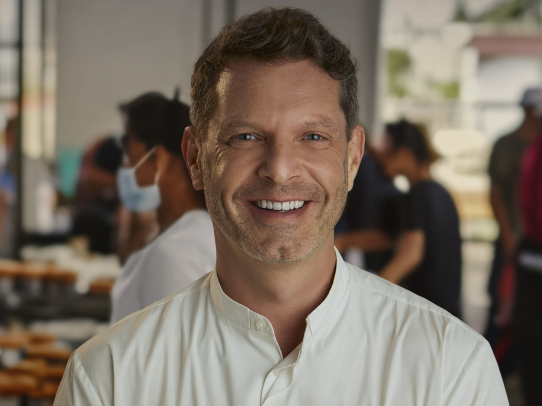 Um homem de meia-idade com barba por fazer e camisa branca sorrindo em um ambiente de trabalho.