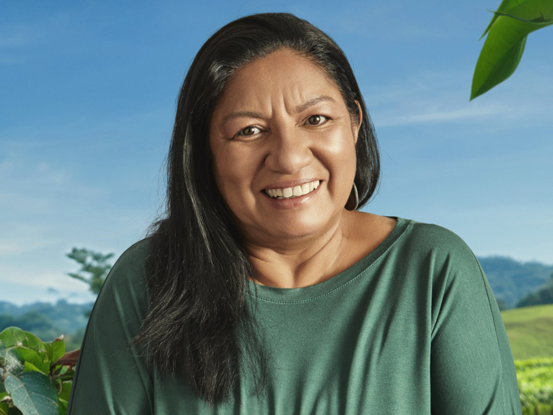 Uma mulher sorrindo, com cabelos escuros e longos, em um cenário de natureza verde.