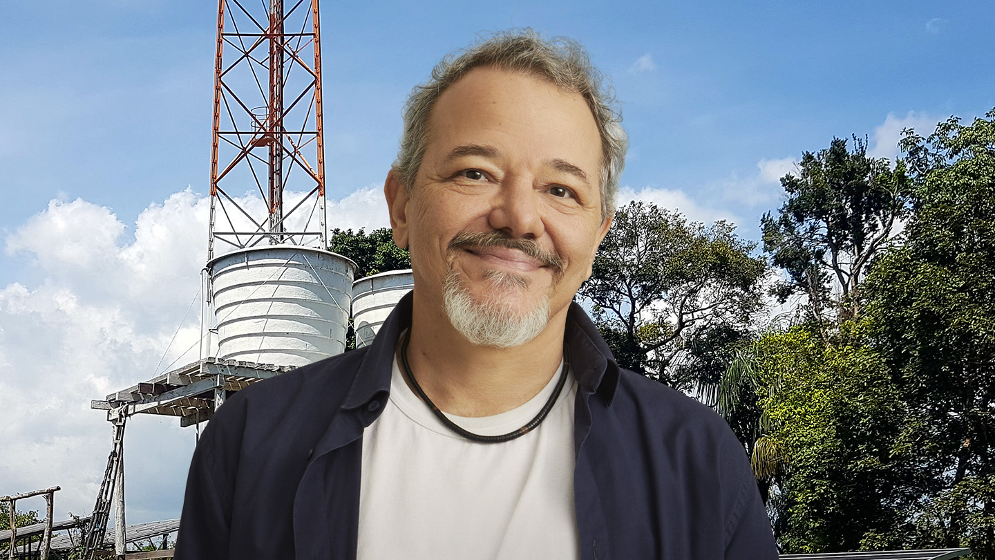 Homem sorridente com barba grisalha, em frente a uma torre de água e árvores.