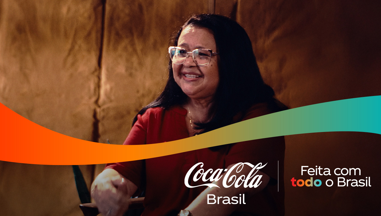 Uma mulher sorrindo, usando óculos e uma blusa vermelha, com o logo da Coca-Cola Brasil e a frase "Feita com todo o Brasil" sobre uma faixa colorida.