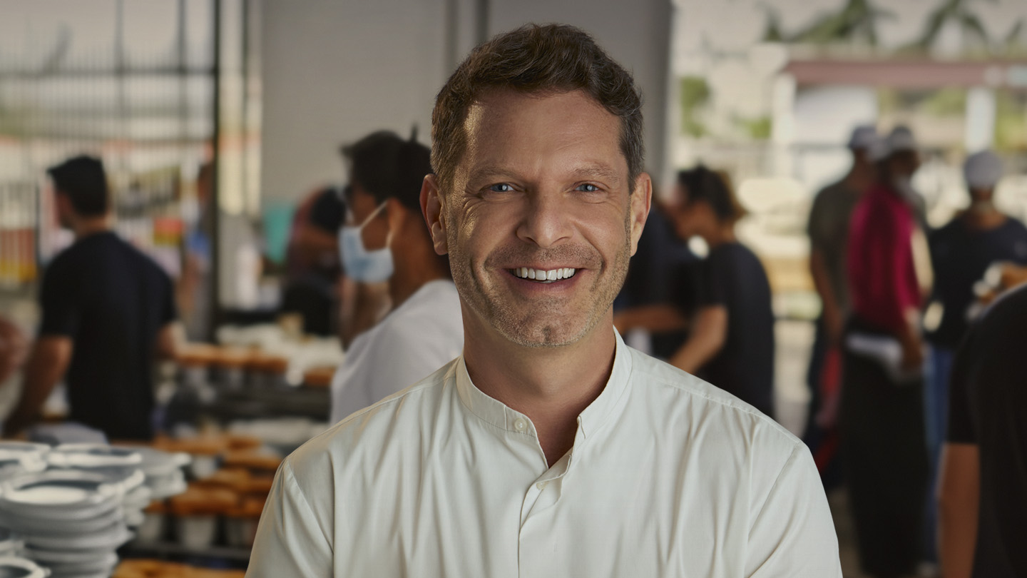 Homem sorridente de camisa branca, com pessoas e pratos ao fundo em ambiente de cozinha ou evento.