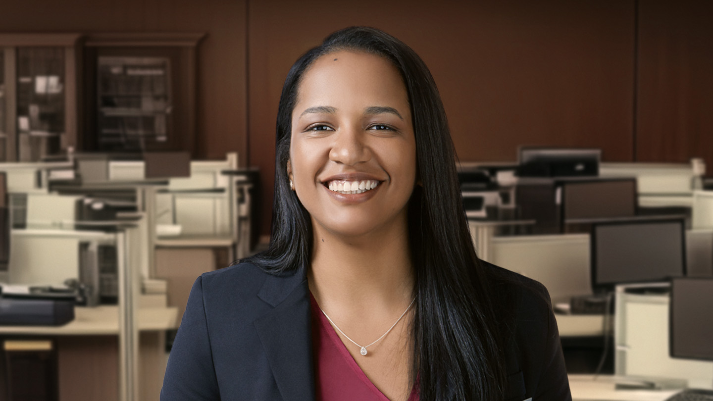 Mulher sorridente com cabelo escuro e colar, em escritório com cubículos e computadores ao fundo.