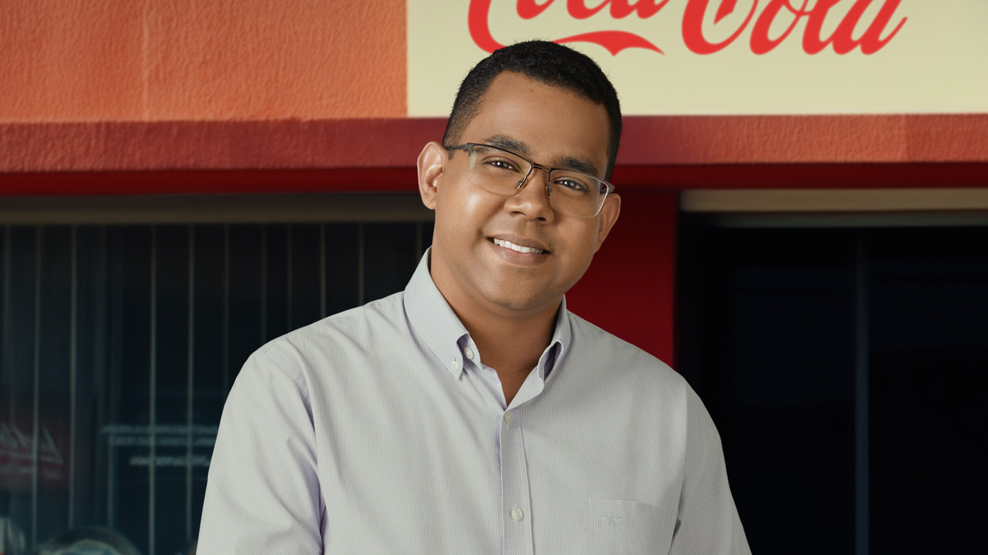 Um jovem sorridente de óculos e camisa clara, em frente a um letreiro vermelho e amarelo da Coca-Cola.