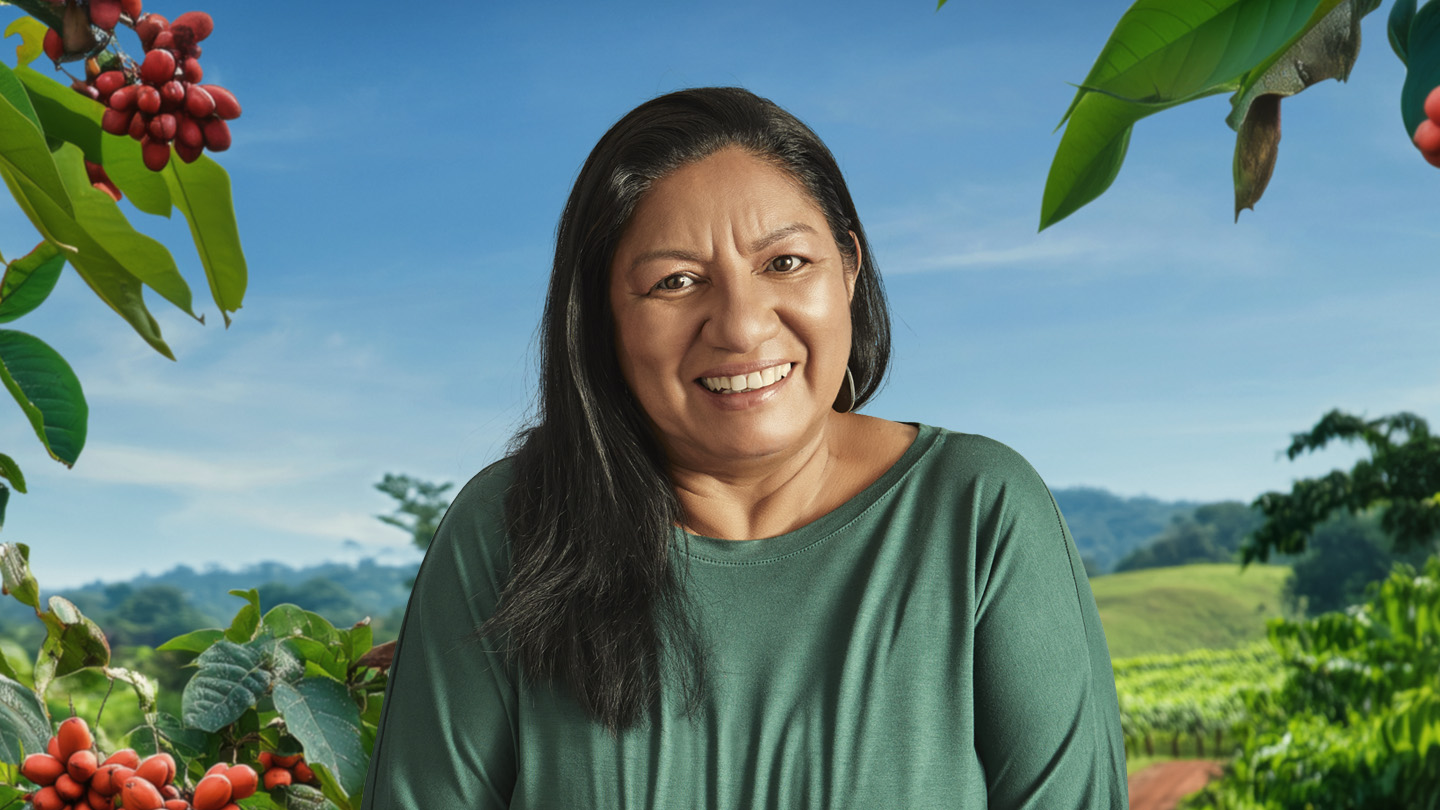 Mulher sorridente com cabelo escuro, em paisagem rural com plantas e céu azul.