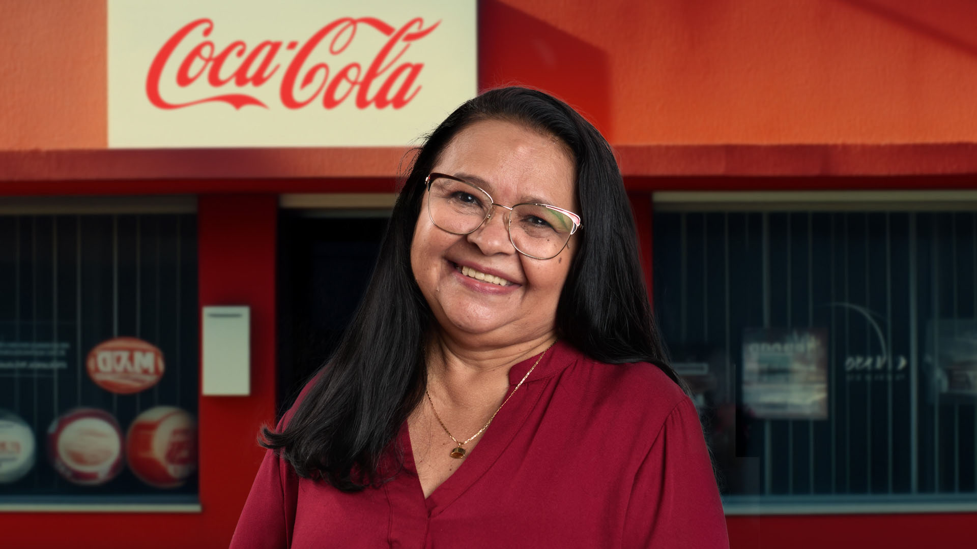 Uma mulher sorrindo, usando óculos e uma blusa vermelha, em frente a uma fachada vermelha com o logo da Coca-Cola.