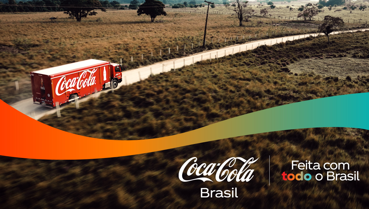 Caminhão Coca-Cola vermelho em estrada rural. Logo Coca-Cola Brasil e "Feita com todo o Brasil" em paisagem seca.