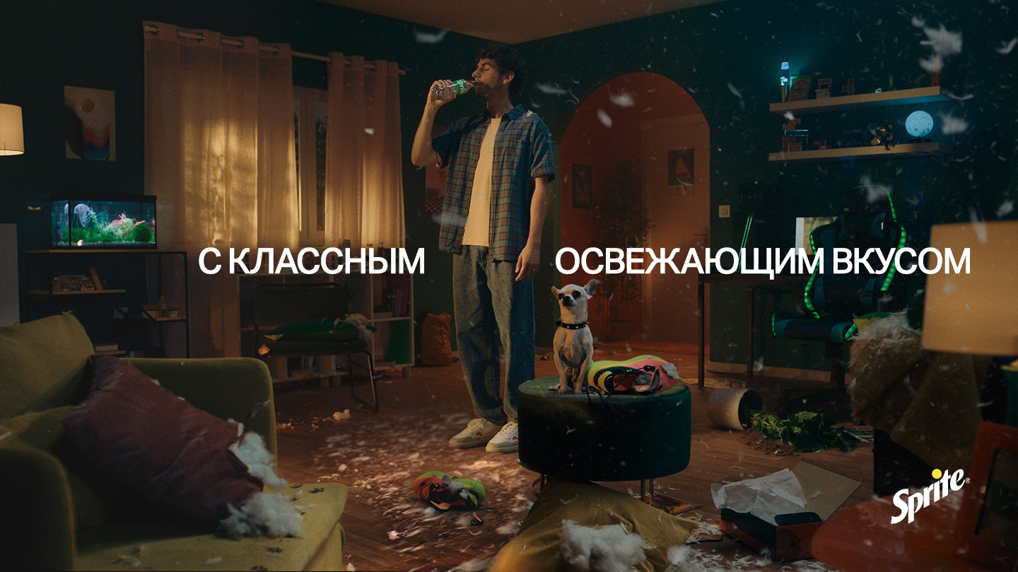 Чалавек п’е Sprite у пакоі з сабакам і разбросанымі падушкамі