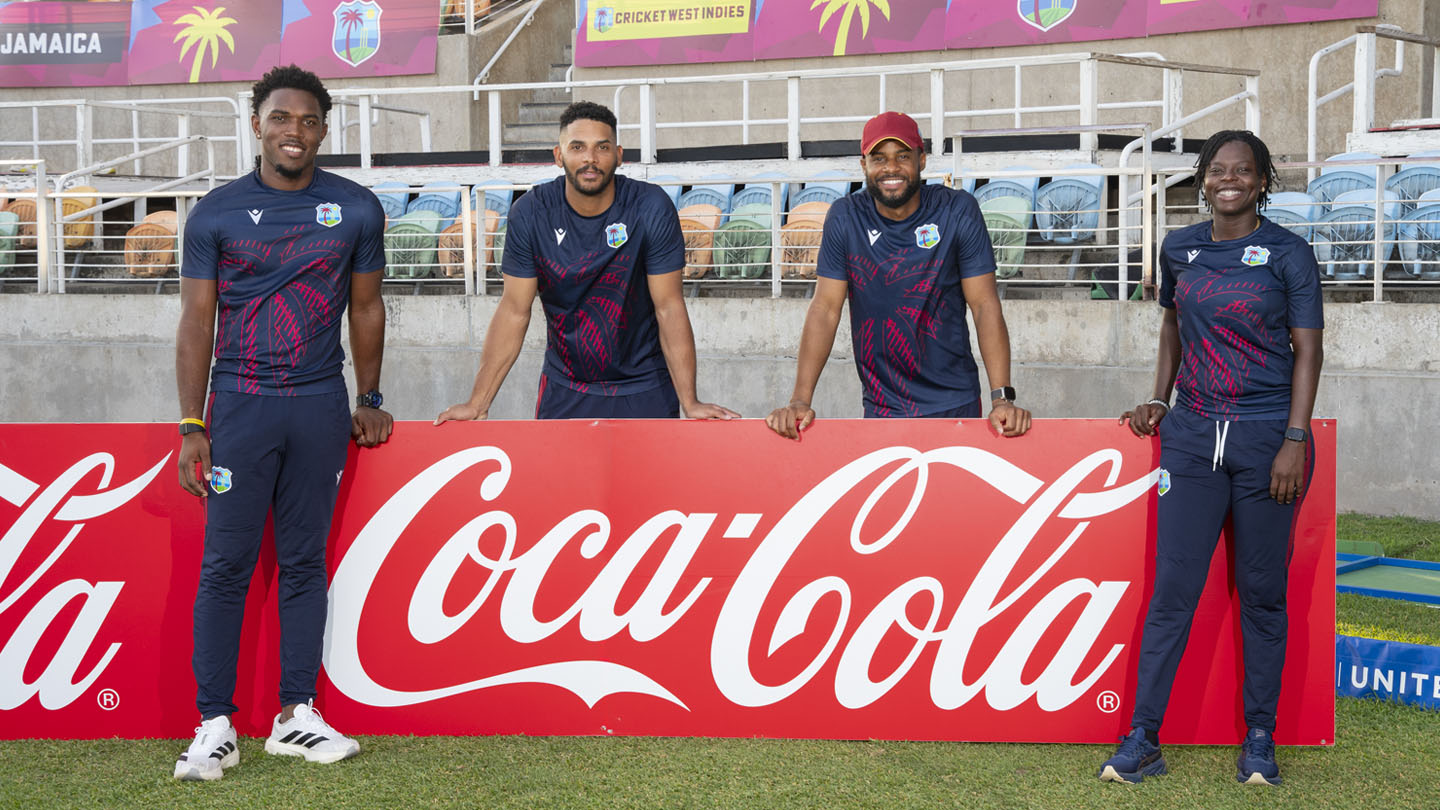 Cuatro jugadores de West Indies Cricket, dos hombres y dos mujeres, posan sonrientes detrás de un gran cartel rojo de Coca-Cola en un estadio.