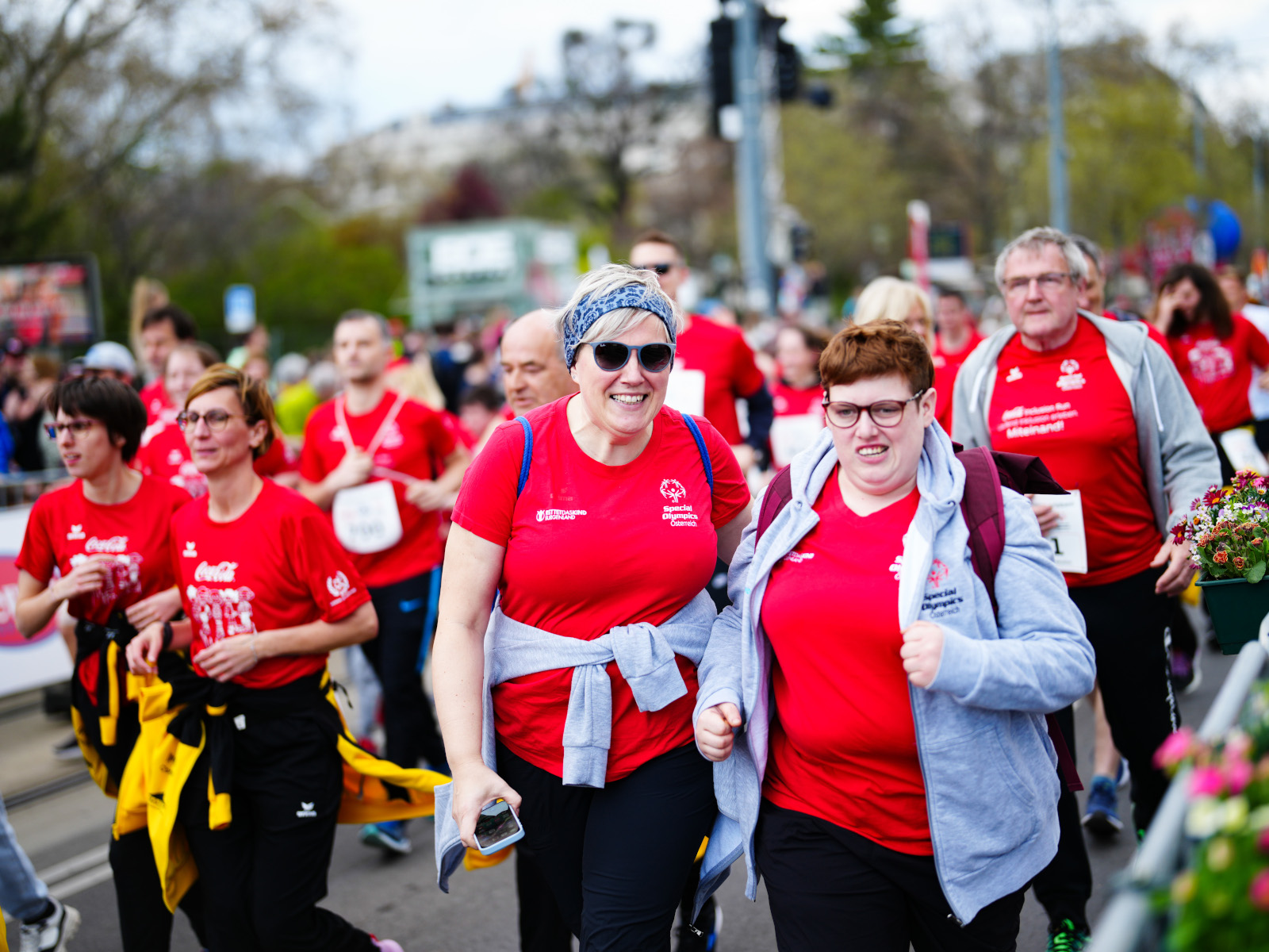 Deux coureuses lors du Coca-Cola Inclusion Run 2025 à Vienne regardent la caméra, tandis que d'autres coureuses parcourent le parcours derrière elles.