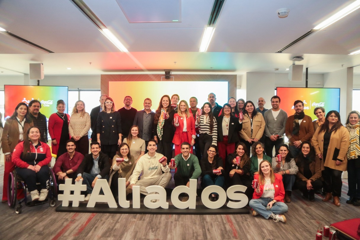 Fotografía de un grupo de personas en una oficina posando junto a un cartel que incluye un hashtag y las letras de la palabra "Aliados"