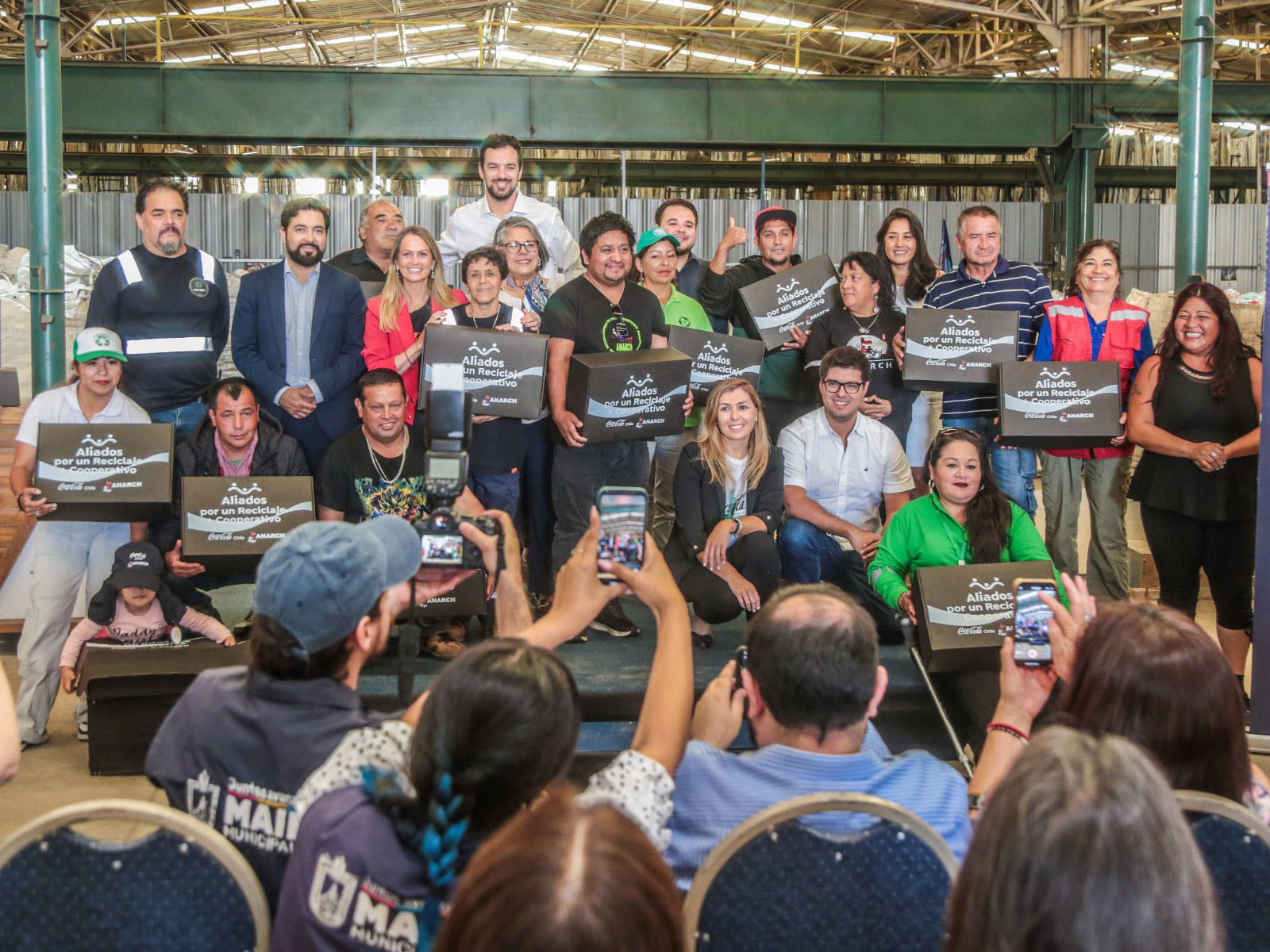 Un grupo de personas son fotografiados sobre un escenario. Algunos sostienen cajas donde se puede leer "Aliados por un Reciclaje + Cooperativo".