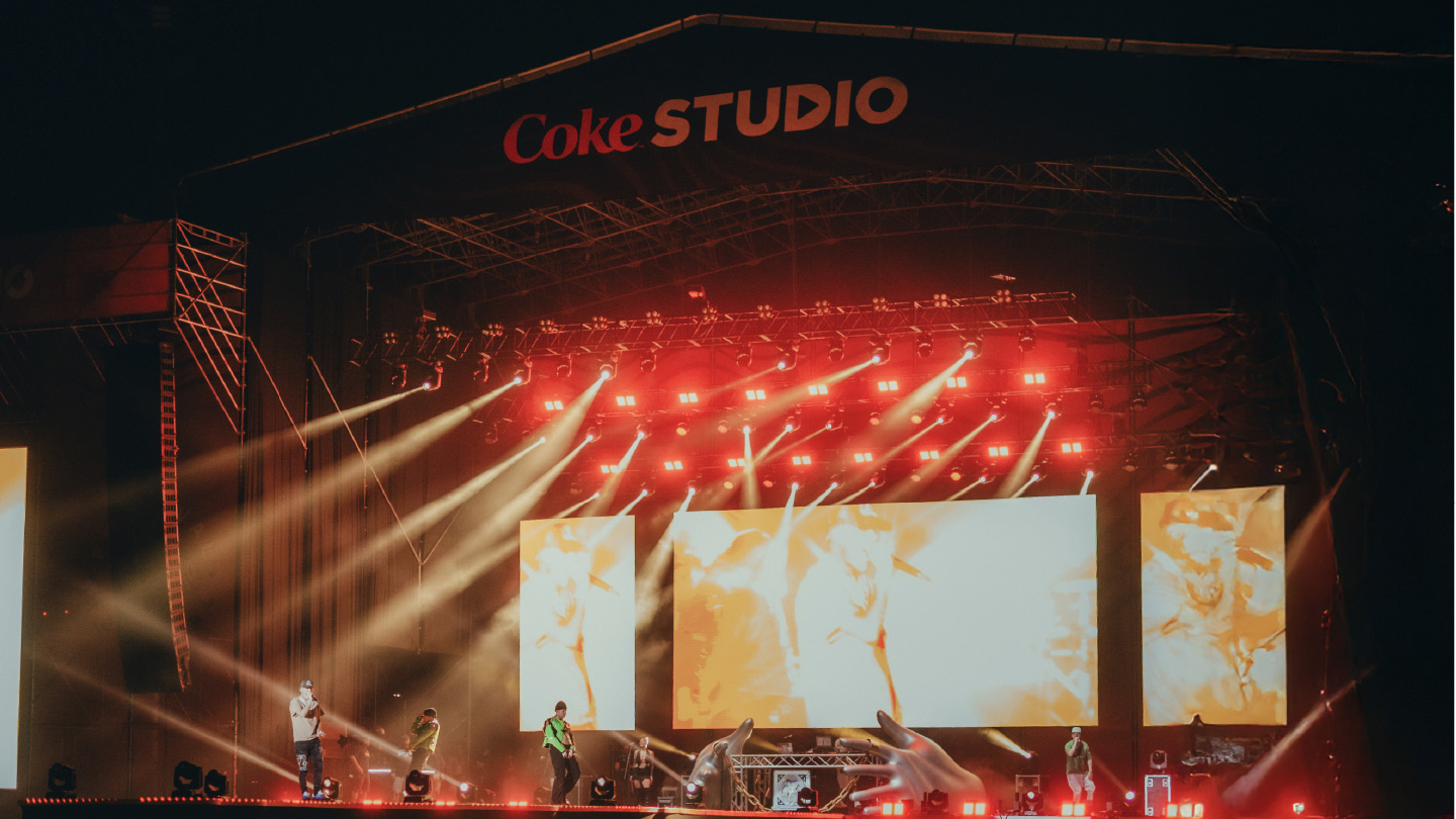 Escenario de concierto de Coke Studio iluminado con luces rojas brillantes y potentes haces de luz, con pantallas grandes mostrando contenido visual. Se aprecian siluetas de personas en el escenario