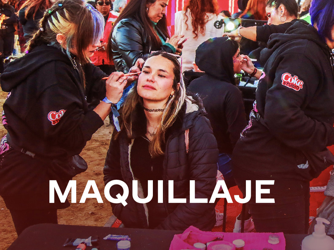 Personas participando en una estación de maquillaje del festival Coke Studio. Los maquilladores, vestidos con sudaderas negras con el logo del evento, aplican diseños creativos a los asistentes. En la parte inferior, aparece el texto "MAQUILLAJE", destacando esta actividad como parte de la experiencia del festival.