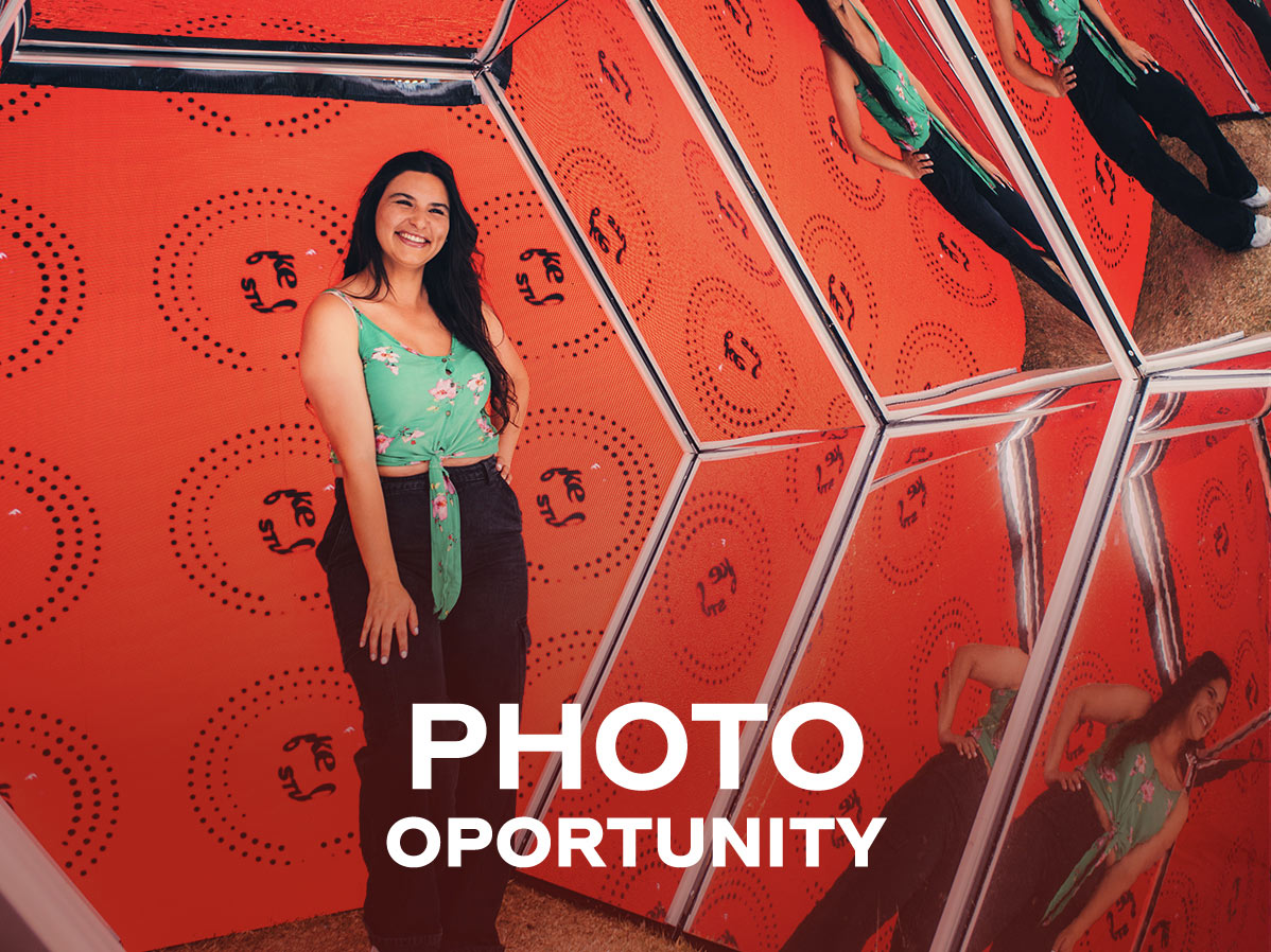 Una persona posando en una instalación interactiva del festival Coke Studio, con un fondo naranja decorado con patrones y logotipos del evento. Las superficies espejadas crean reflejos llamativos que realzan la experiencia visual. En la parte inferior, el texto "PHOTO OPPORTUNITY" destaca esta actividad como un espacio ideal para fotografías creativas.