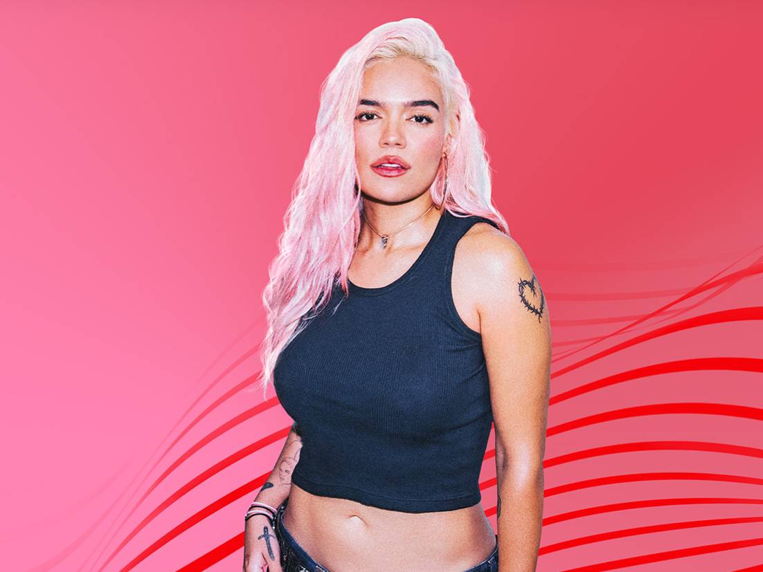 Karol G posando con cabello rosa y ropa casual, frente a un fondo rosa vibrante.