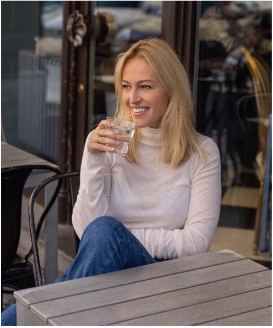 Žena sedí u stolku v kavárně a drží sklenici vody.