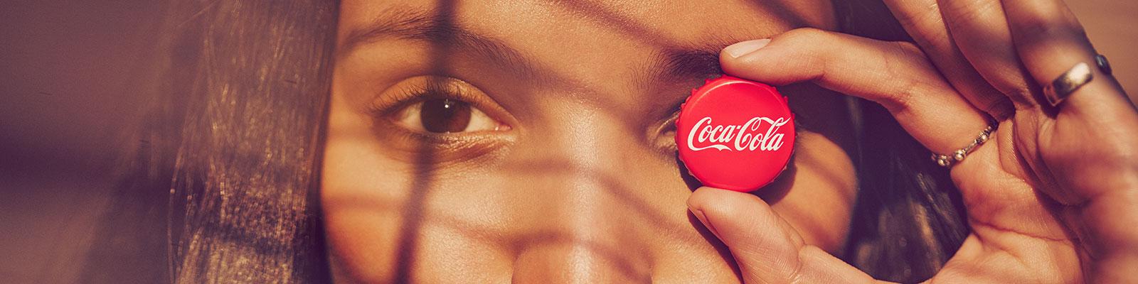 Käsi hoiab punast Coca-Cola pudelikorki silma ees, valgus ja varjud langevad üle kujutise.