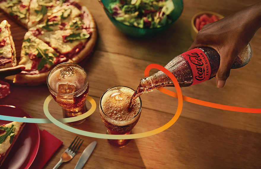 Käsi valab Coca‑Cola pudelist jooki klaasi, laual on pitsaviilud ja salat.