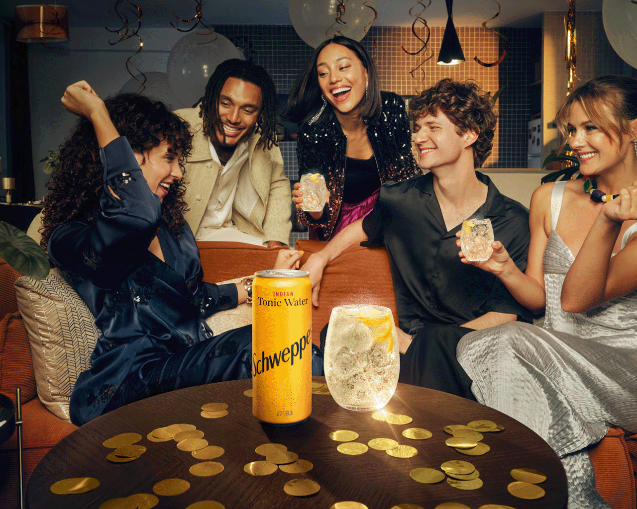 Skupina prijateljev praznuje ob mizi, polni zlatih konfeti. Smejijo se in nazdravljajo s kozarci tonika Schweppes ter pločevinko Schweppes Indian Tonic Water v ospredju. V ozadju so baloni in praznično vzdušje.