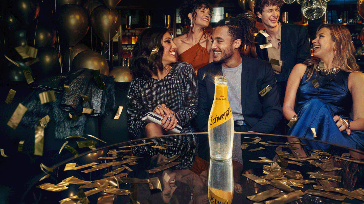 Skupina prijateljev, elegantno oblečenih, se smeji in uživa v baru. Na mizi je steklenica Schweppes tonika, ki je v ospredju slike. Vzdušje je praznično in sproščeno.