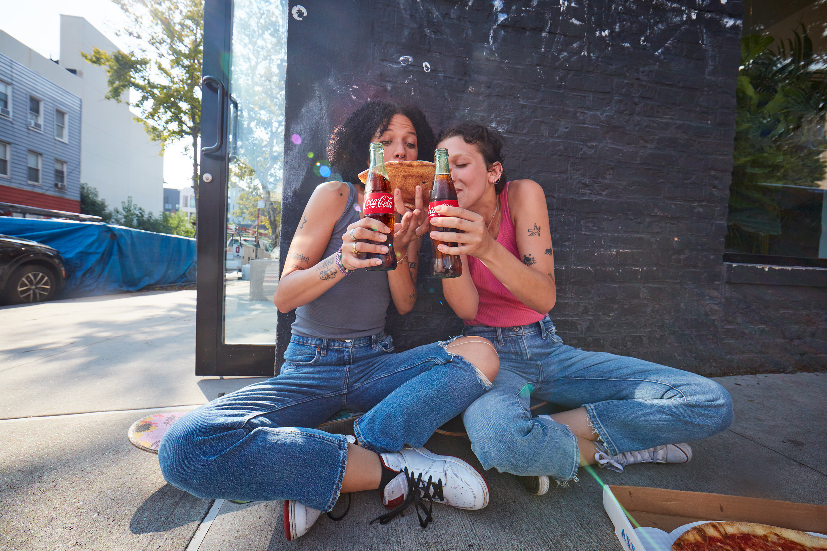 Foto met twee personen zittend op een stoep, houdend Coca‑Cola flessen en delend een pizzapunt.